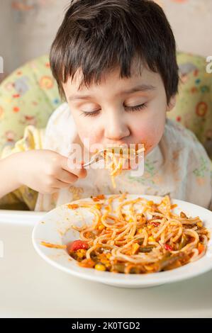 Bambino che mangia spaghetti con verdure. Bambino che mangia divertente. Ragazzo dai capelli castani con il viso coperto di salsa. Fine settimana, ambiente caldo e accogliente in cucina Foto Stock