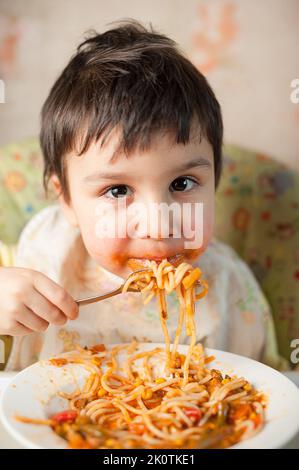 Bambino che mangia spaghetti con verdure. Bambino che mangia divertente. Ragazzo dai capelli castani con il viso coperto di salsa. Fine settimana, ambiente caldo e accogliente in cucina Foto Stock