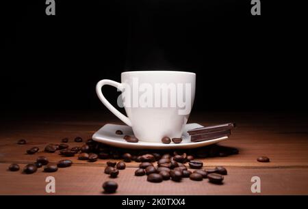 Piccola tazza di caffè caldo, pezzi di deliziosa tavoletta di cioccolato e chicchi di caffè appena macinati sparsi su un tavolo di legno, con sfondo nero. Foto Stock