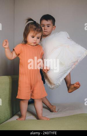 Felice ragazzo allegro giocando in salotto con la sua sorella bambino che lancia cuscini in piedi sul divano a casa. I bambini felici godono di passatempo attivo, spenda Foto Stock