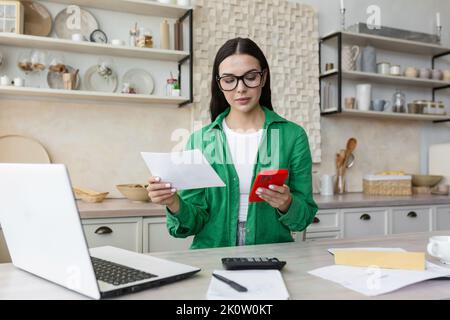 Lavoro a casa. Una bella giovane commercialista lavora a casa in cucina, si siede al tavolo con un computer portatile e una calcolatrice. Tiene in mano documenti e un telefono rosso. Foto Stock