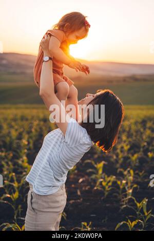 Madre che alza sua figlia quando si trova in piedi in mezzo ai campi di mais verde in una giornata di sole estate mentre si cammina sulla natura all'aperto. Famiglia felice Foto Stock