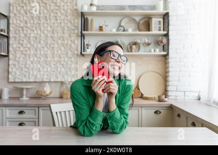 In amore e felice giovane bella donna invia e riceve messaggi d'amore sul telefono rosso da una persona amata, da un ragazzo. In piedi in cucina a casa in bicchieri e una camicia verde. Foto Stock