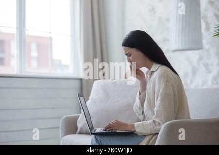 Quarantena. Guerra. Una bella giovane donna si siede a casa sul divano con un notebook, parla online tramite video link con la sua famiglia e gli amici, grida, non li sente. Pulisce le lacrime con un tovagliolo. Foto Stock