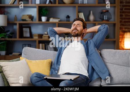 Bell'uomo che lavora sul computer portatile mentre si siede sul divano in un appartamento elegante. Rilassati dopo il lavoro. Sollevato dopo il lavoro, mettere le mani dietro la testa e. Foto Stock