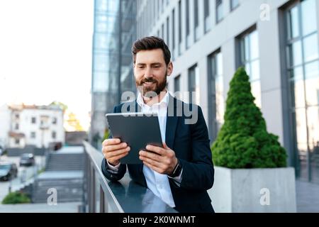 Allegro sicuro attraente giovane uomo caucasico con barba in tuta chattando su tablet, ha riunione all'aperto Foto Stock