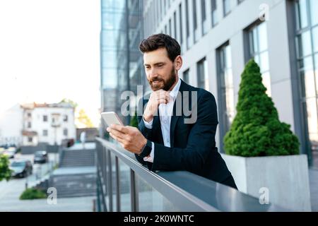 Giovane uomo caucasico, sorridente e gentile, con barba in tuta, orologio su smartphone sul balcone Foto Stock