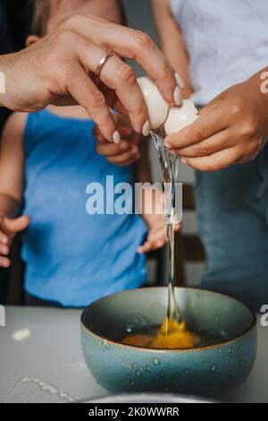Mani della madre che mostrano ai suoi bambini come rompere le uova fresche in una ciotola per omelette mentre preparano gli alimenti insieme nella cucina. Famiglia felice. Divertimento per tutta la famiglia. Foto Stock