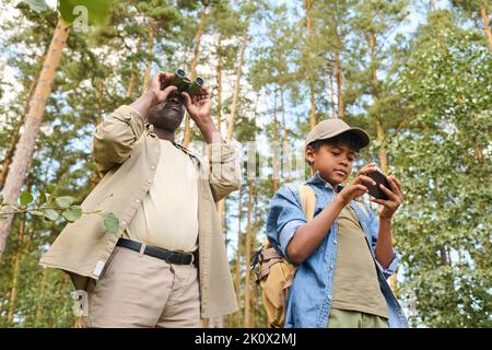 Bambino con zaino e smartphone che utilizza la mappa del navigatore durante il viaggio nella foresta mentre si trova contro il nonno con binocolo Foto Stock