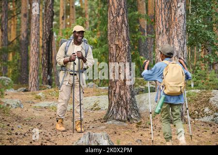 Felice uomo anziano con bastoni da trekking in piedi sul sentiero nella foresta e guardando il suo nipote che si muove verso di lui durante l'escursione Foto Stock