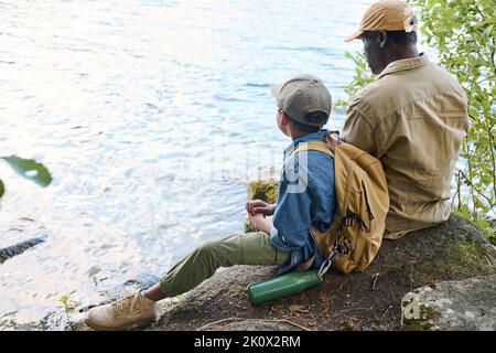 Vista posteriore del ragazzo afroamericano con zaino e suo nonno seduti sul mare e riposarsi mentre si gode un'escursione nel fine settimana Foto Stock