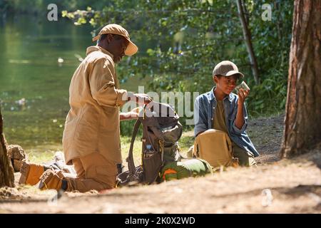 Un anziano uomo afroamericano che disimballa lo zaino mentre si trova in ginocchio sul sentiero davanti al nipote con un vasetto di cavallette Foto Stock