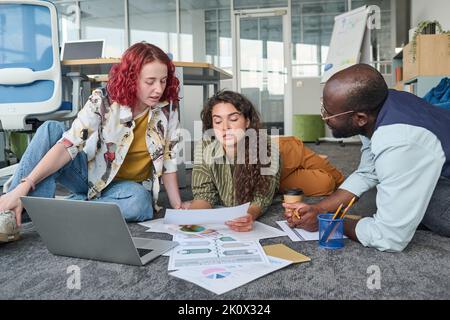 Giovane afro-americano che ascolta colleghi di sesso femminile che discutono di documenti finanziari sdraiati sul pavimento dell'ufficio di fronte al computer portatile Foto Stock