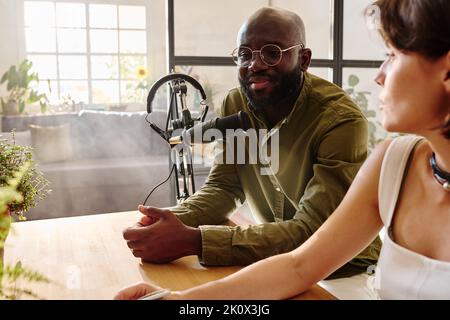Giovane ospite afro-americano maschio e donna bianca che hanno conversazione su argomento d'attualità e registrazione di file audio per il pubblico online Foto Stock