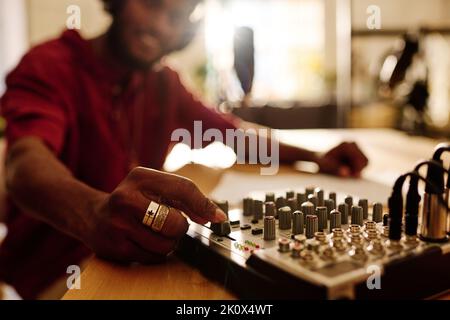 Mano di un giovane uomo che ruota la console del mixer mentre si siede sul posto di lavoro in studio e regola il livello del suono prima di registrare un nuovo file audio Foto Stock