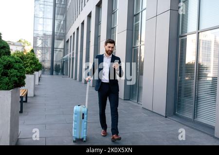 Felice fiducioso giovane maschio caucasico con barba in tuta con valigia va, chattando da smartphone Foto Stock