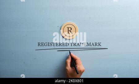 La lettera R è tagliata in cerchio di legno tagliato e la mano maschile scrive un marchio registrato sotto di esso. Su sfondo di legno blu pastello. Foto Stock