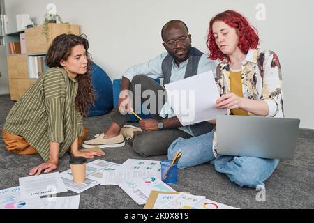 Una giovane economista che spiega i dati contenuti nel documento finanziario ai colleghi multiculturali seduti sul pavimento accanto a loro Foto Stock