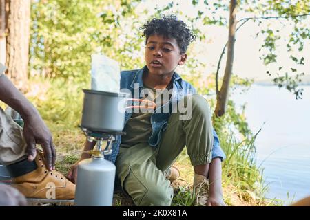 Carino afro-americano pre-teen ragazzo che parla con il nonno mentre si siede di fronte a lui e padella con cibo durante il fine settimana escursione Foto Stock