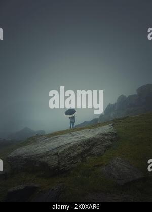 La vista posteriore di un uomo solitario con ombrello si erge su una collina rocciosa coperta da foschia. Moody e scena emotiva con una silhouette unica estranea sotto la ra Foto Stock