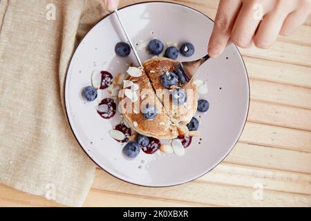 La donna tiene posate mentre fa colazione - frittelle tradizionali con mirtilli freschi e sciroppo di agave su un piatto. Prima colazione all'aperto sul terr Foto Stock