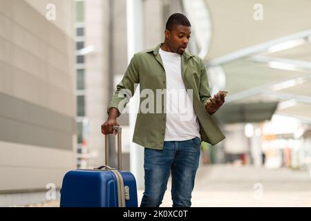 Scioccato il ragazzo nero usando il telefono in piedi con la custodia da viaggio all'aperto Foto Stock