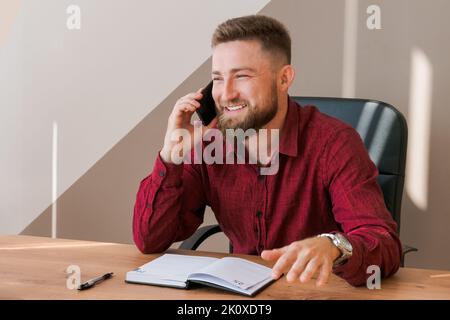 Un uomo d'affari concentrato e bearded nei vestiti dell'ufficio seduti al tavolo e parlando sullo smartphone, notando le informazioni in un blocco note. Il concetto di negoziazione e pianificazione di un business di successo Foto Stock