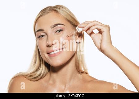 Sorridente bella giovane donna che applica siero, olio sul viso su sfondo bianco. Concetto di cosmetici, trucco, bellezza naturale Foto Stock