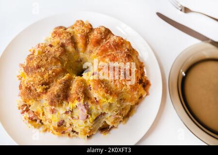 Crostata salata al forno su piatto. Pasta bollita con uova miste e un pezzo di carne affumicata cotta in teglia su piatto, posate e piatto vuoto su fondo bianco Foto Stock