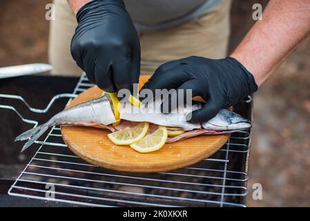 Sgombro alla griglia con fettine di limone, vista dall'alto. Il processo di cottura del pesce all'aperto. Chef in guanti in lattice nero preparazione di sgombro pesce su un bosco Foto Stock