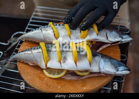 Chef in guanti in lattice nero prepara sgombri di pesce su un tagliere di legno, processo di spruzzatura di spezie e sale Foto Stock