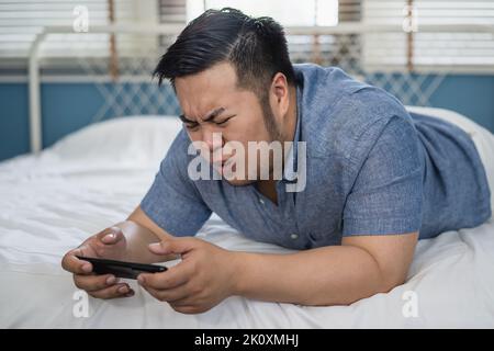 Giovane asiatico più grande uomo che gioca un videogioco sul telefono cellulare e ha ottenuto reazioni negative a letto a casa. Le espressioni facciali potrebbero essere furiose, feroci, sconvolgite, infelici, irritate, frustrato. Foto Stock