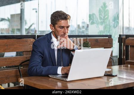 Imprenditore maschile pensivo in tuta toccando mento e pensando ai dati su netbook mentre si siede a tavolo e lavorando su progetto di business in caffè Foto Stock