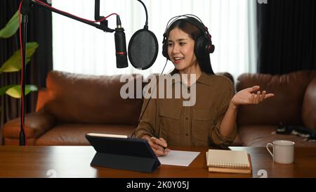 Giovane donna che usa il microfono a condensatore e le cuffie per registrare podcast in un piccolo studio domestico Foto Stock
