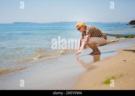 Donna anziana attraente che tocca l'acqua sulla riva del mare Foto Stock