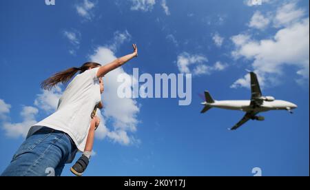 Vista dal basso della donna con il bambino che agita la mano al volo aereo commerciale nel cielo. Stile di vita e concetto di viaggio. Foto Stock