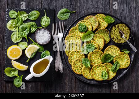 frittelle di spinaci su piatto nero con pesto di basilico e ingredienti su tavola di pietra su tavolo di legno scuro, vista orizzontale dall'alto, piatto Foto Stock