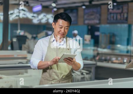 Addetto alle vendite asiatico del negozio di alimentari in grembiule utilizzando tablet digitali contare le merci nel dipendente del supermercato che esegue l'ispezione di prodotti inventario regolare per controllare le merci ordine online Foto Stock