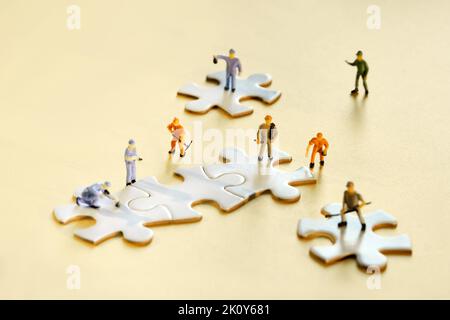 Squadra di minuscoli lavoratori in miniatura su pezzi di puzzle collegati jigsaw su carta giallo dorato. Primo piano su elementi puzzle bianchi. Foto Stock