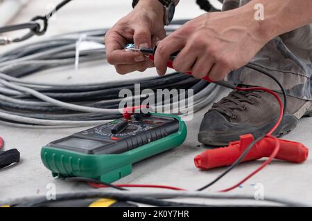 Primo piano della mano di un ingegnere elettrico che suona un cavo con un multimetro o che misura la tensione in un cantiere. Lavoro di ingegneria debole. Ingegnere, cavo, multimetro, segnale di linea, tensione, mani, dita, primo piano, cantiere, produzione, tecnologia, elettronica Foto Stock