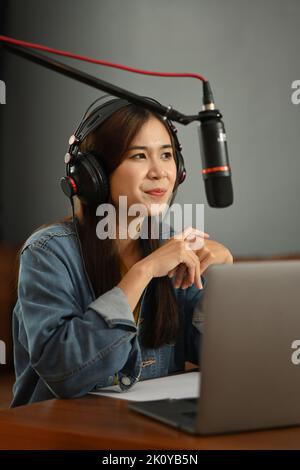 Ospite radio donna positivo che utilizza il microfono condensatore e il laptop per registrare podcast in un piccolo studio domestico Foto Stock