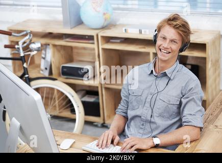 Ascoltare musica in ufficio. Ritratto di un giovane designer che usa il suo computer mentre è seduto in ufficio. Foto Stock