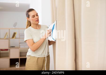 Giovane donna che stira le tende a casa Foto Stock