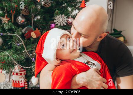 Papà bacia suo figlio, vestito in costume da Babbo Natale, vicino all'albero di Natale. Foto Stock