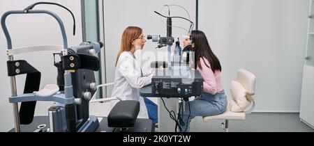L'optometrista caucasico che esegue il test visivo per la donna giapponese presso una clinica oftalmologica moderna. Esame oculare e diagnostica visiva Foto Stock