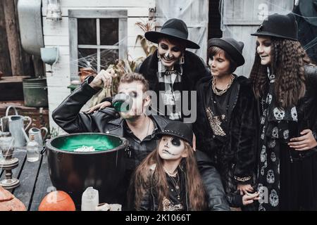 Famiglia spaventosa, madre, padre, figlie che celebrano Halloween. Cauldron di potion.Terrificante nero cranio mezzo-faccia trucco, costumi strega, elegante immagine Foto Stock