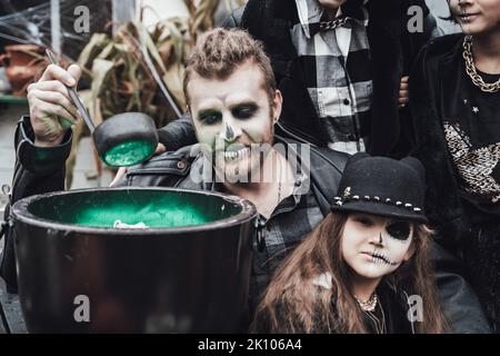 Famiglia spaventosa, madre, padre, figlie che celebrano Halloween. Cauldron di potion.Terrificante nero cranio mezzo-faccia trucco, costumi strega, elegante immagine Foto Stock