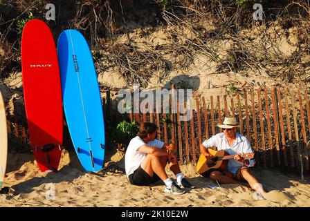 Due uomini adulti cantano e suonano la chitarra sulla spiaggia con le loro tavole da surf dietro loro in un giorno di sole vacanze estive Foto Stock
