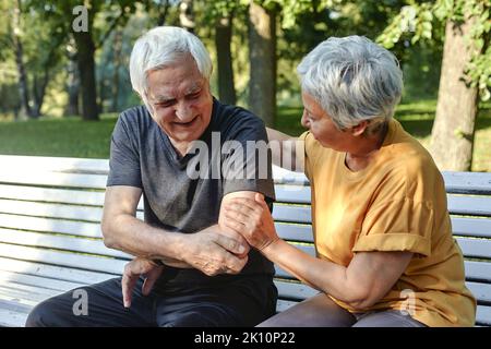 Durante la passeggiata sportiva mattutina o facendo esercizi nel parco, l'anziano 70s uomo si ferì la spalla o il gomito che afferrava il braccio seduto in panca con la cura di Foto Stock