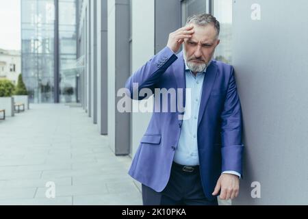 L'uomo anziano, l'operaio, l'uomo d'affari in una tuta si trova vicino al centro dell'ufficio, si appoggia contro il muro, tiene la mano dietro la testa, sente un mal di testa. Nervi, stress sul lavoro. Foto Stock